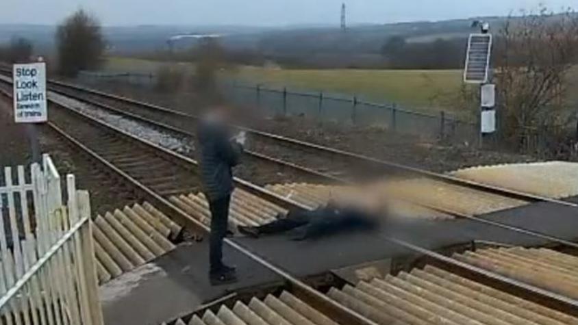 A person wearing black is lying down on the railway track. Their face is blurred. Another person in black is standing and taking a photo on a phone.