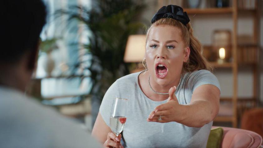 Polly is holding a champagne flute in her hand and engaged in what is clearly a heated discussion with her mouth open and her empty hand outstretched towards a person she is talking to but who we can only see blurred and from the back