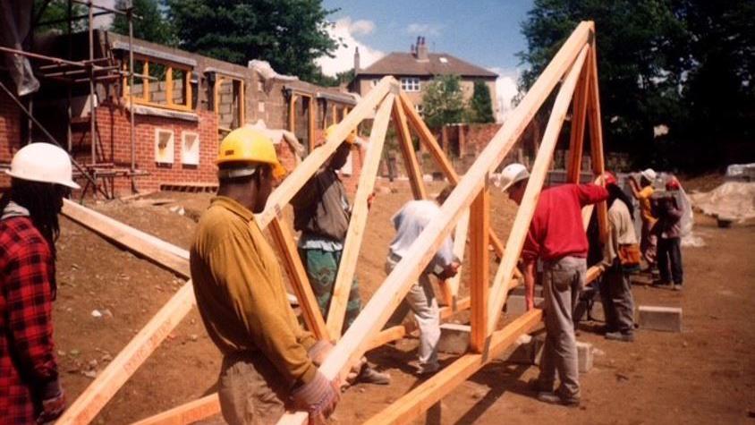 An old image of people carrying a section of roofing in Leeds. In the background is a housing construction site. The people are wearing hard hats.