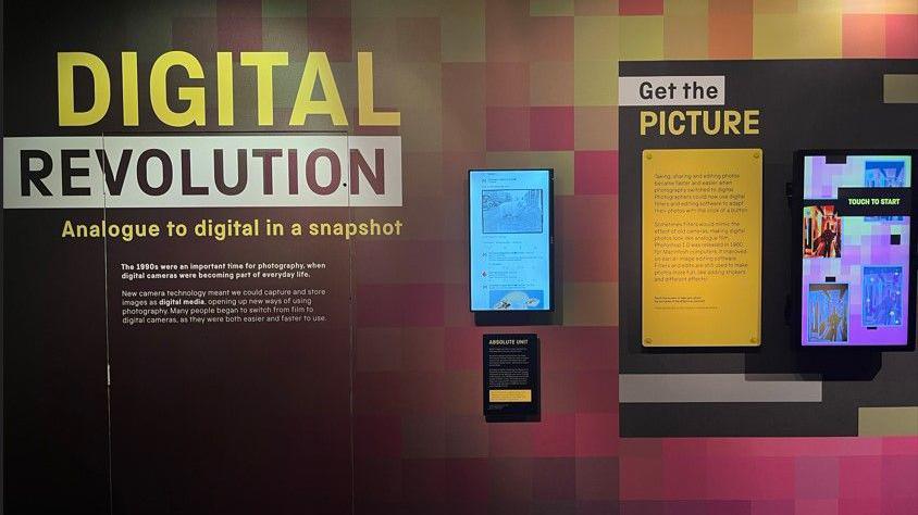 An interactive display screen shows the meme, with information boards around it. The board says "Digital revolution. Analogue to digital in a snapshot." There is also a sign saying "absolute unit" with information below it, and another sign saying "get the picture" with writing below.