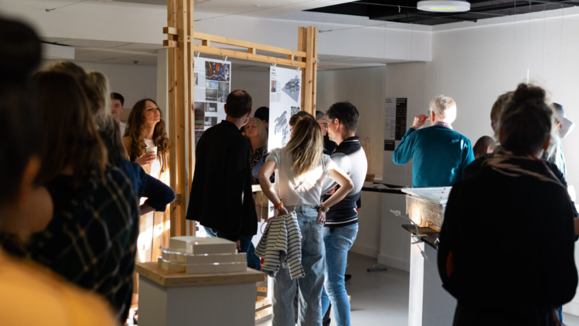 A group of people gather around a wooden frame, bulletin board type structure with information on it at a history exhibition in a light, airy room.