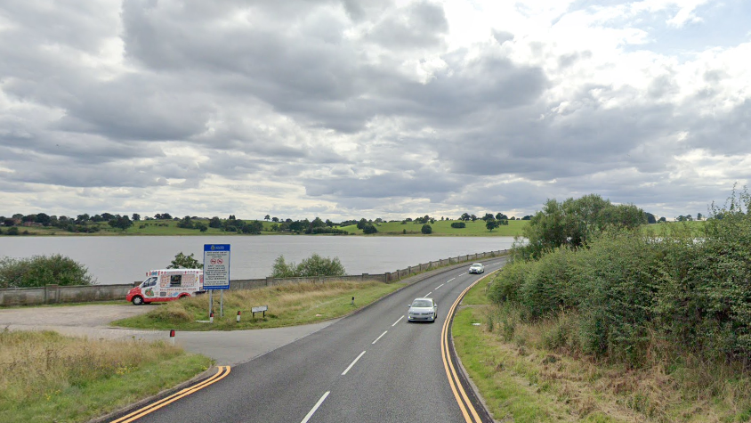 A road with a curved bend. On its left is a small car park with an ice cream van parked up. Beyond that is a large reservoir with green fields and trees around it.