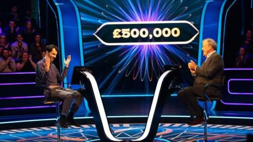 Two men sitting down opposite each other. The man on the left is wearing brown trousers and a brown shirt and has his hands in the air. The man on the right is wearing a brown suit and clapping. There is a sign with £500,000 in white lettering in the background. There are people sat down on either side clapping.