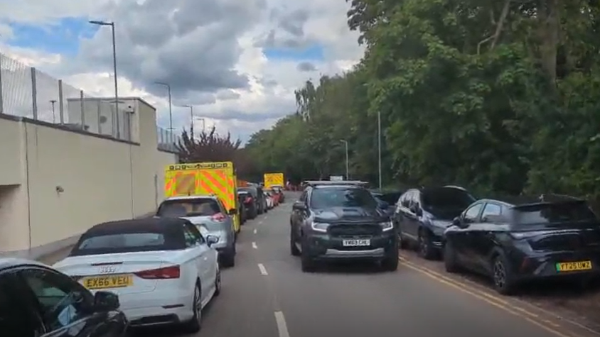Cars and ambulances wait in a long queue, with one car travelling without traffic in the opposite direction. The street is tree-lined.