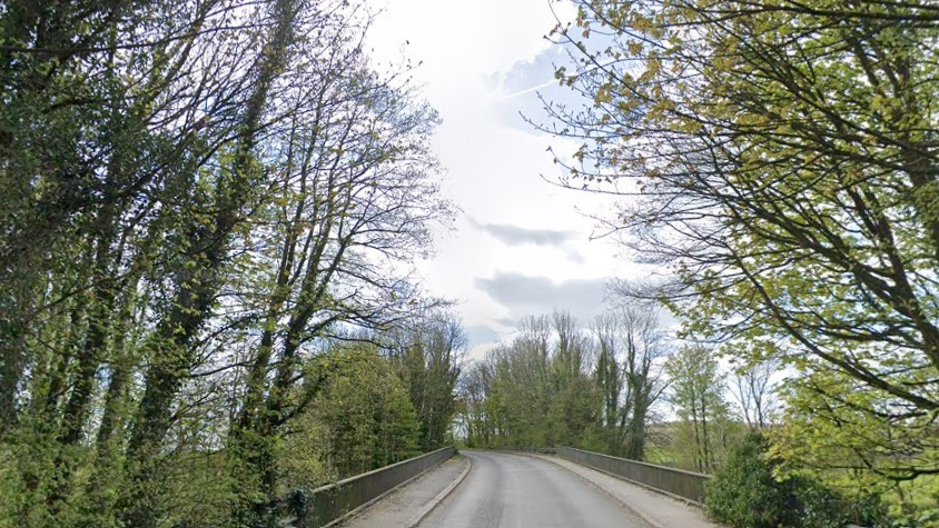 Brigsteer Bridge on Brigsteer Road near Kendal. The road is lined with green trees. The bridge has grey metal railings on either side.