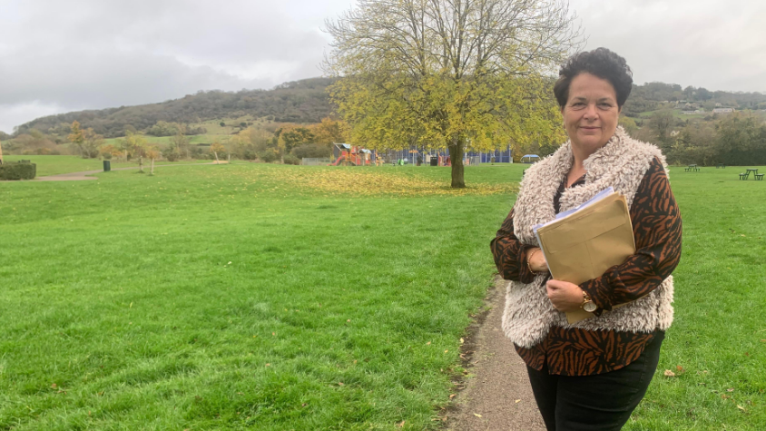 Zoey Holland stands in a large green field with a park in the background. A tree with lime leaves blocks the full park from view. Zoey stands in the foreground in a furry waistcoat and holding a brown paper file.