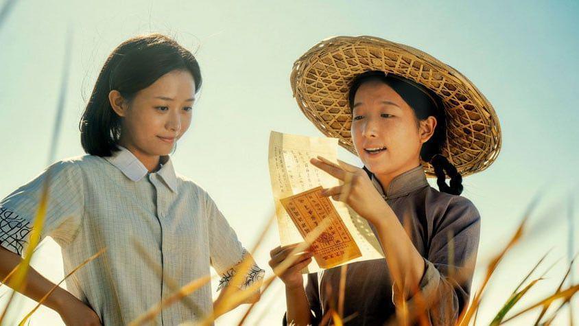 Two women standing in a field, one is reading a letter out to the other.