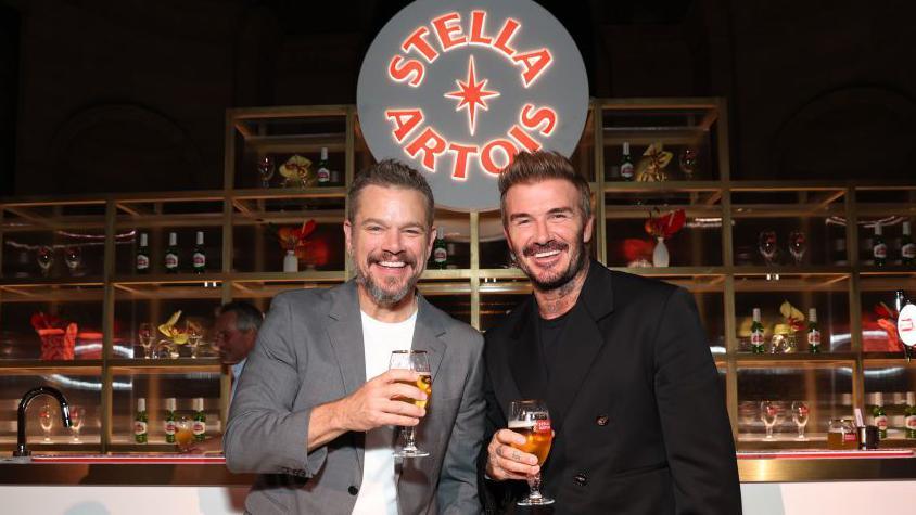 Matt Damon and David Beckham stand next to each other in front of a bar while holding glasses of Stella Artois, a sign for the beer brand can be seen behind them.