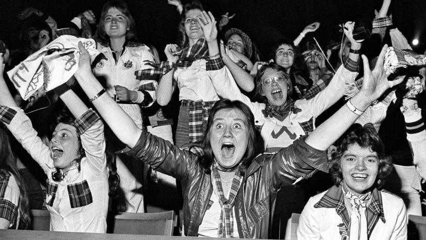 A crowd of Roller fans scream in delight as their idols perform on stage, all wearing bits of tartan with their hands in the air. The girl at the front is wide-eyed and open-mouthed in excitement as she raises her hands in the air.