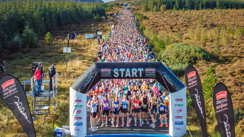 hundreds of runners behind a start gate on a road through a forest