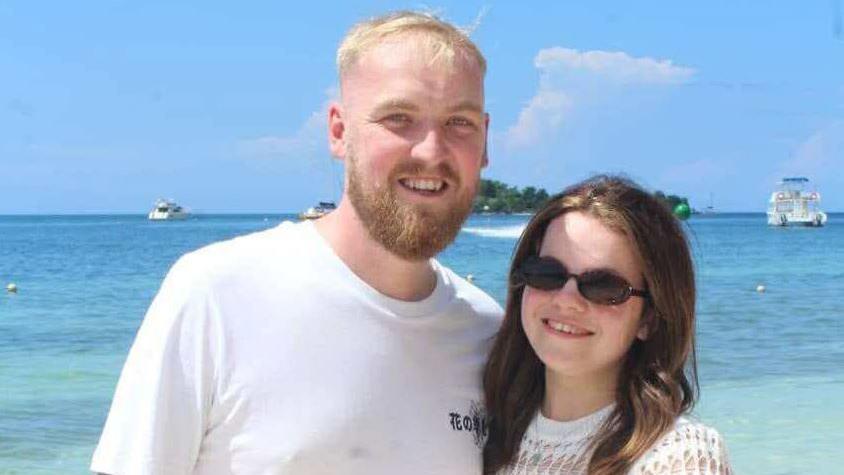 A man with short blond hair and a beard wearing a white T-shirt is next to a shorter woman with long brown hair and a white top. They are standing in front of a blue sea with boats, with a small island in the distance.