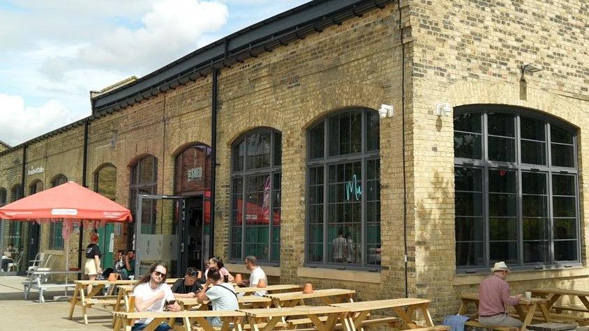 The shed is an old brick building with lots of benches and seats outside. It has very large arched windows and people are sitting outside enjoying the sunshine