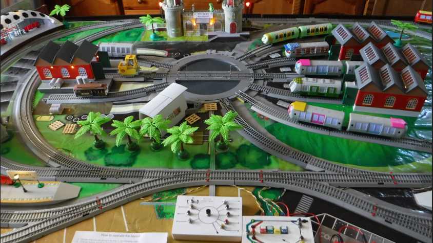 A model railway layout with several trains emerging from buildings on a figure of eight track and two control boards in the foreground 