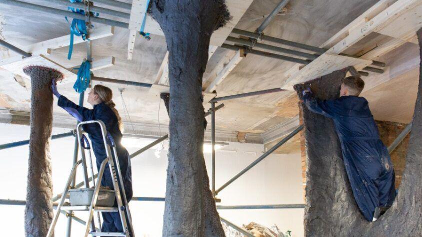 Knitted structure hanging from a ceiling with people in boiler suits layering brown paste on to it. 