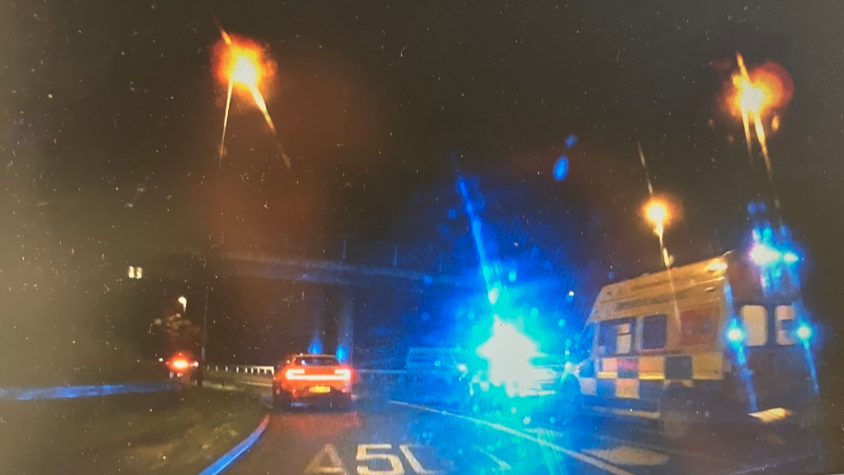 Ambulances and a police car are pulled up on a lane of a dual carriageway. It is dark, and their blue lights are reflecting off the camera