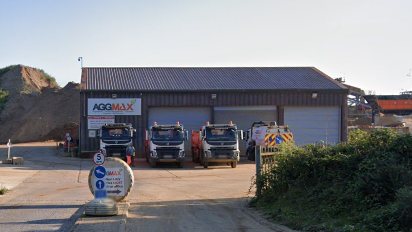 An entrance to a quarry. A building can be seen with four HGV style vehicle parked outside of it in a row. A large mound of earth can be seen to the left of the building.