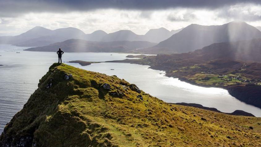 There is a long piece of grass with a man standing at the end. There is the sea below and mountains in the distance.