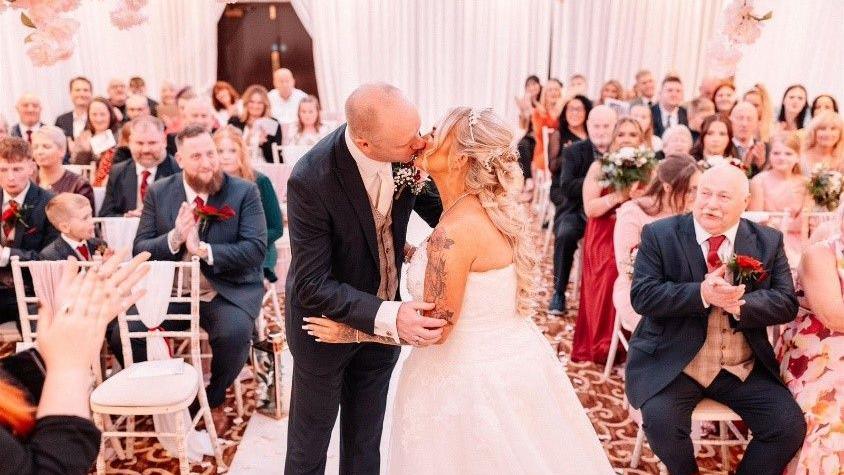 Lisa Marsay, who has long blonde hair and tattoos on her arm, kisses her husband while wearing a white wedding dress, stood in the middle of a wedding aisle with guests sat either side clapping. 