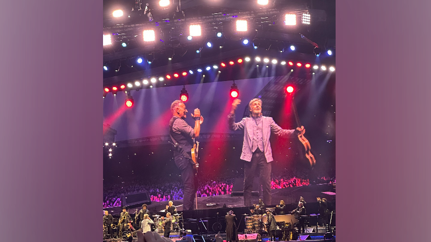 A big screen and stage, showing Sir Paul McCartney holding his hands up to the crowd and holding a guitar. Bruce Springsteen is standing to his left clapping, holding a guitar and smiling. There is red and blue lighting on stage with other musicians.