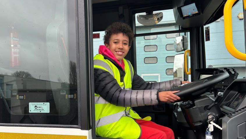 A child sit in the sit of a bin lorry with his hands on the steering wheel, he is wearing a hi-visibility vest over a black puffer jacket and red trousers