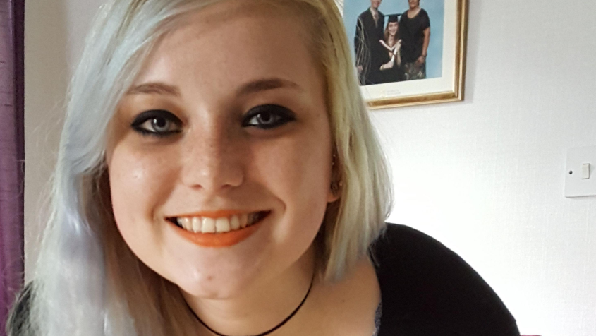 A smiling Rebekah Morris with dyed blonde hair in front of a picture from her graduation ceremony