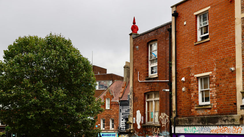 A red sculpture of a boy wearing a long pointed 'dunce' hat can be seen perched on the edge of a city building, with the figure's back facing the camera. The picture is taken from a distance, and red-brick buildings can be seen in the foreground and background. A large tree is visible on the left of the image. 