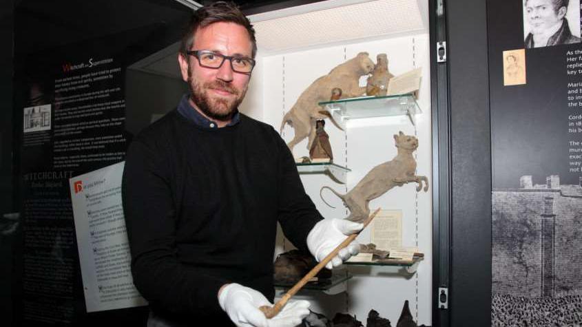 Daniel Clarke smiles at the camera holding a witches wand in front of an exhibition. He has short dark hair and he wears black glasses with a black jumper. He wears white gloves.