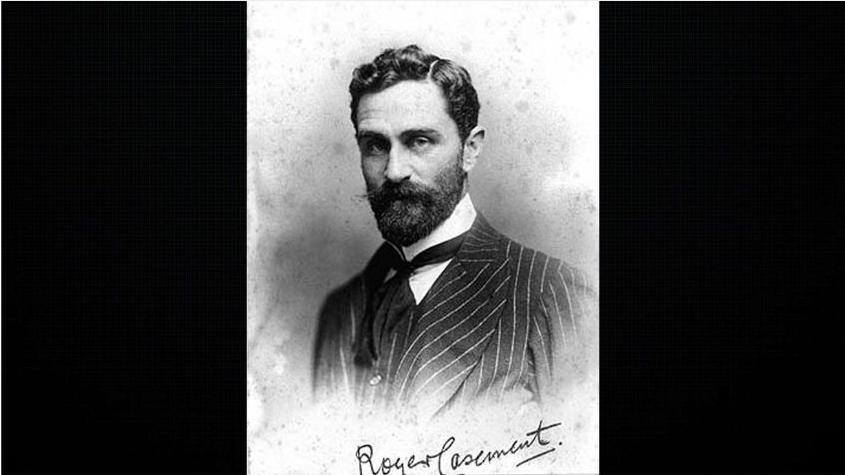 A black and white portrait of Roger Casement wearing a pin strip suit, white shirt and dark cravat. 