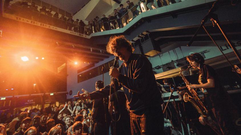 A band member of Fat Dog is singing into a microphone to a crowd that is around him. There is a woman playing the saxophone behind him. Everything is lit up in a golden light.