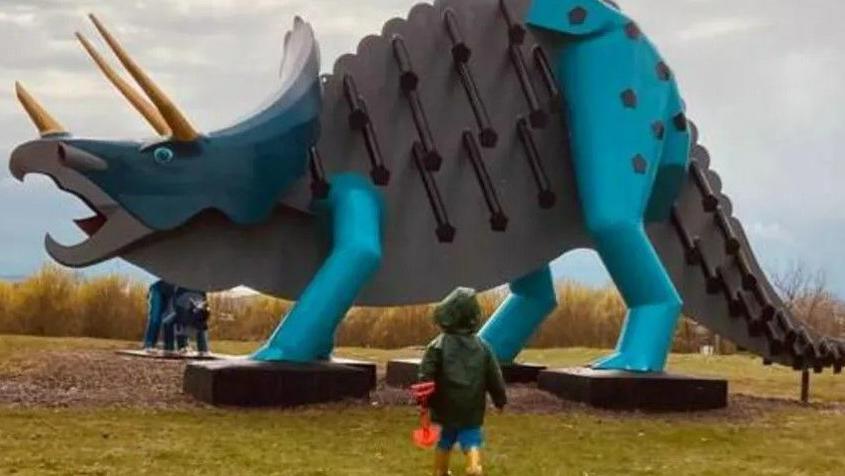 A small child in a green raincoat and yellow wellies is pictured from behind carrying a red spade and walking towards a steel triceratops that towers above him. A smaller dinosaur is visible in the background.