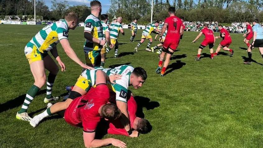 Players from Guernsey Raiders and Jersey RFC are playing rugby on a grass pitch. In the foreground three men are on the floor.