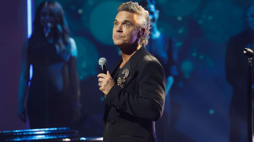 Robbie Williams wearing a black suit with an ornate brooch while holding a microphone, which suggests he is performing on stage. He had a grey mullet/mohawk hairstyle.