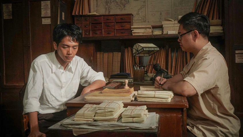 A man sits across another man at a desk. One of them is writing on a piece of paper.