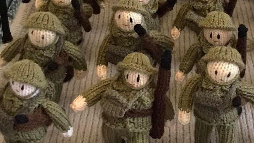 Six soldiers, created from yarn. They are wearing amy uniform and carrying packs and guns.