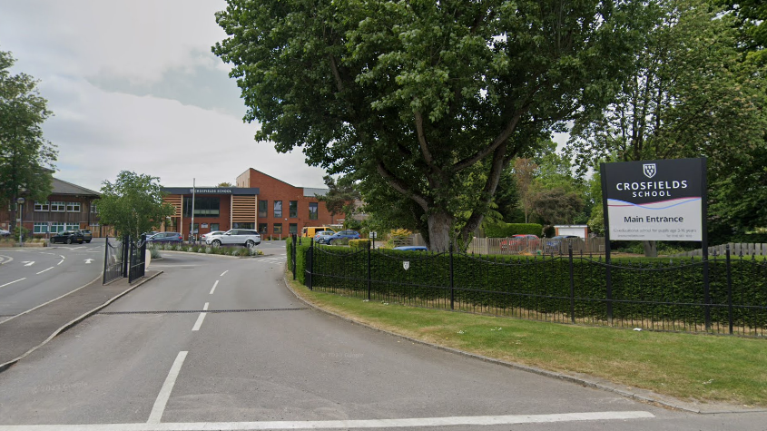 The entrance to a school, which has a navy and white sign outside reading "Crosfields School Main Entrance". It is fenced off with a gate. Beyond the gate are a number of brick school buildings.