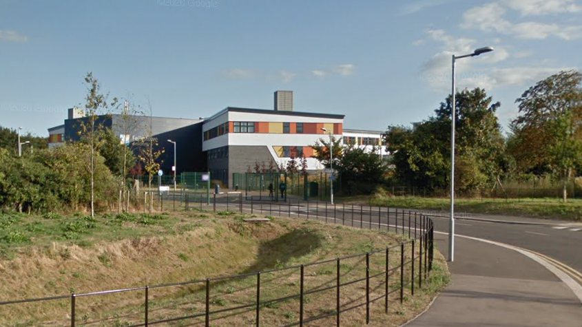 The Felixstowe School building can be seen in the background while a road and greenery can be seen in the foreground. The building is comprised of two square blocks - one dark grey, the other white with orange, yellow and read panes. 