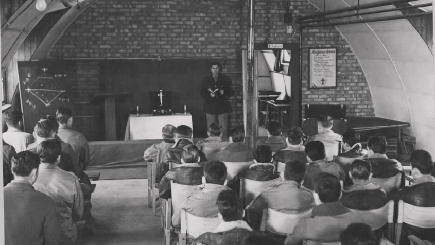 A black and white photo of rows of men sitting on chairs. In front of them is a table with a cross on it. A man at the front is holding a Bible while addressing the congregation.