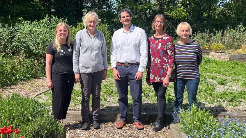 Four women and one man stand in a row and pose for a photo in a green area, surrounded by plants and bushes.