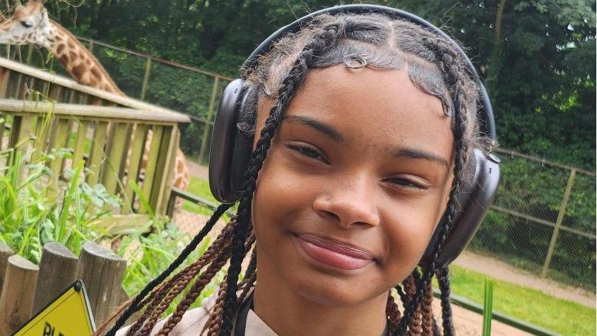 A teenage girl wearing headphones smiles at the camera in front of a giraffe enclosure; a giraffe can be seen in the top left corner