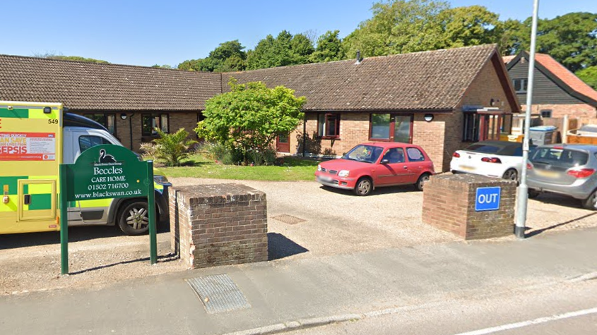 A Google Street View image of the entrance to Beccles Care Home - a small brick building with a dark brown roof. Cars are parked outside and an ambulance can be seen on the left of the image.