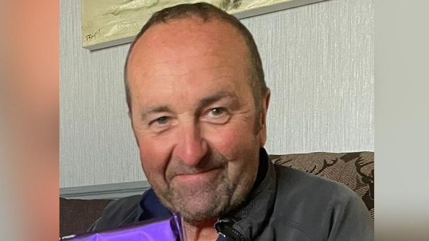 David Burrows smiles at the camera, sitting on a sofa with part of a painting visible on the wall behind him. Wearing a grey zipped jacket and a dark top, he appears to be holding a present wrapped in shiny purple paper.