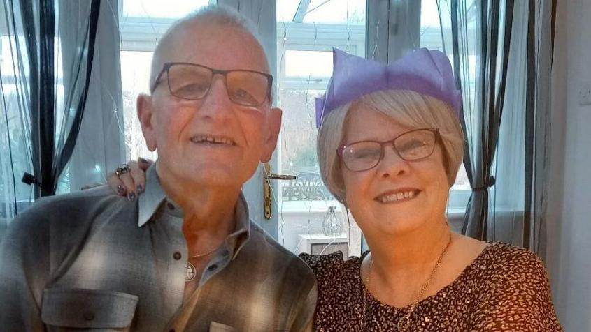 A couple pose with their arms around each other and smile into the camera. A woman, on the right of the image, is wearing a purple paper Christmas hat and a brown patterned top. A man on the left is wearing a grey and white checked shirt. They are both wearing spectacles.
