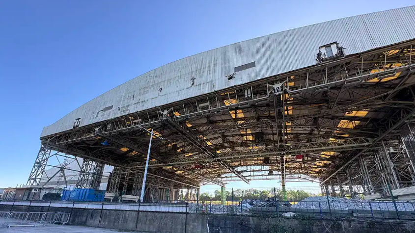 A large warehouse building which is in the process of being demolished. It has a grey corrugated-style roof still intact but much of the internal part of the building has been removed to the point where just the frame is visible. The picture is taken on a clear, sunny day with blue sky overhead