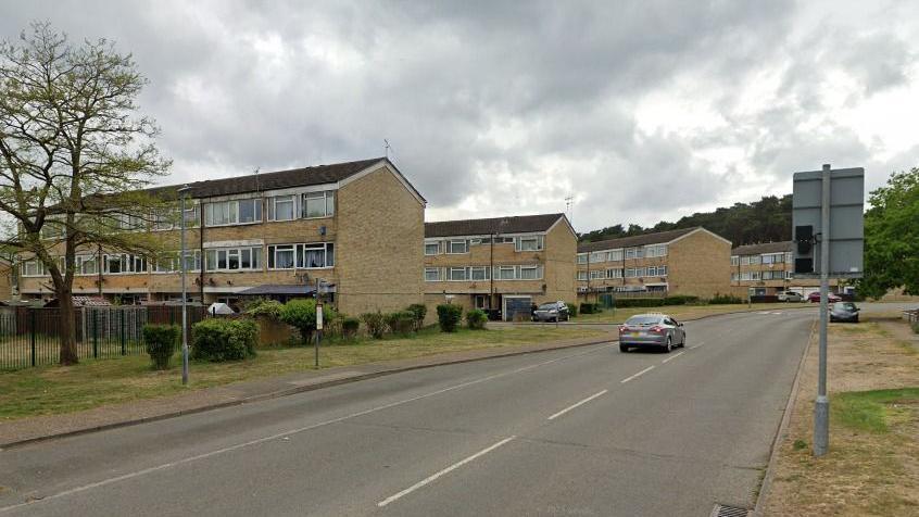 A large housing estate with four-two-story blocks. Cars can be seen on the road and in the distance. There are grass verges with shrubbery and trees in the background.