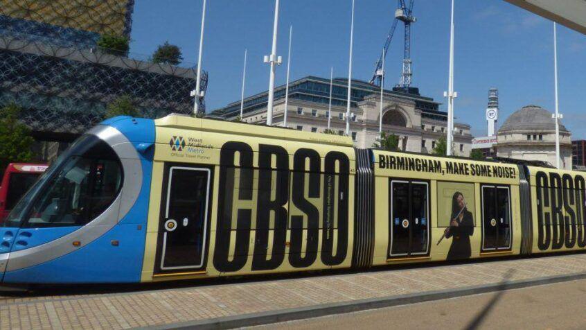 A blue and yellow tram in a city. It is decorated with black writing that says "CBSO Birmingham, make some noise!" there is a photo on the tram of a woman playing the flute