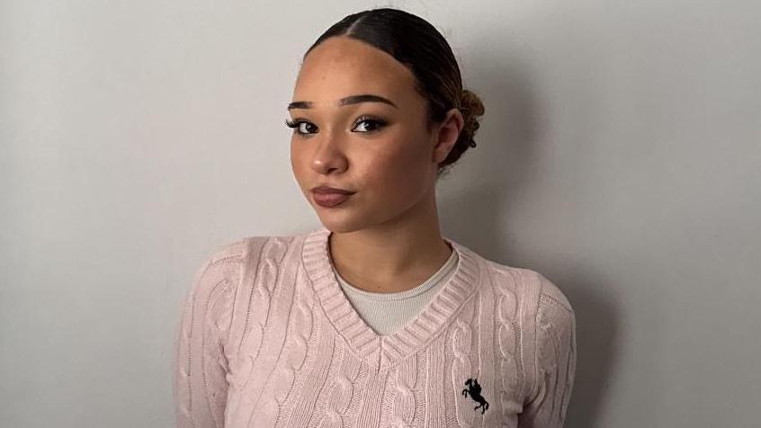 Demi, 17 is smiling at the camera, wearing a pink v-neck sweater and has her hair in a ballet bun.