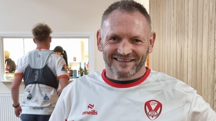 A smiling Gary McKee in a visitor centre wearing a red and white top and inside a community centre where behind him you can see an open hatch 