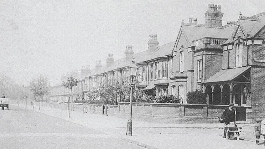 Old black and white photo of Carholme Road with the house in the corner and a man with a wheelbarrow and another man with a trailer in the distance