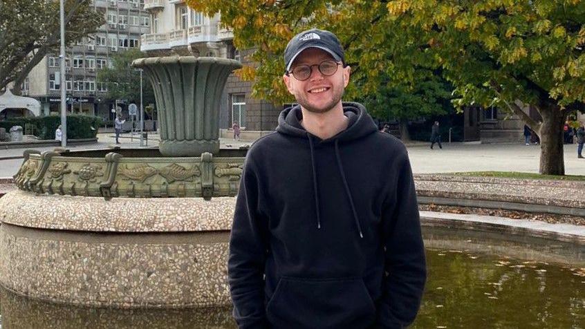 A man wearing a black hoodie stands by a fountain. He is also wearing glasses and a black baseball cap. There are trees in the background and other people can be seen walking around a paved open space.