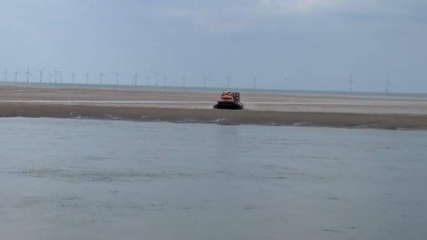 A yellow RNLI hovercraft can be seen in the distance on the Leasowe sands with water in the foreground and wind turbines in the background.